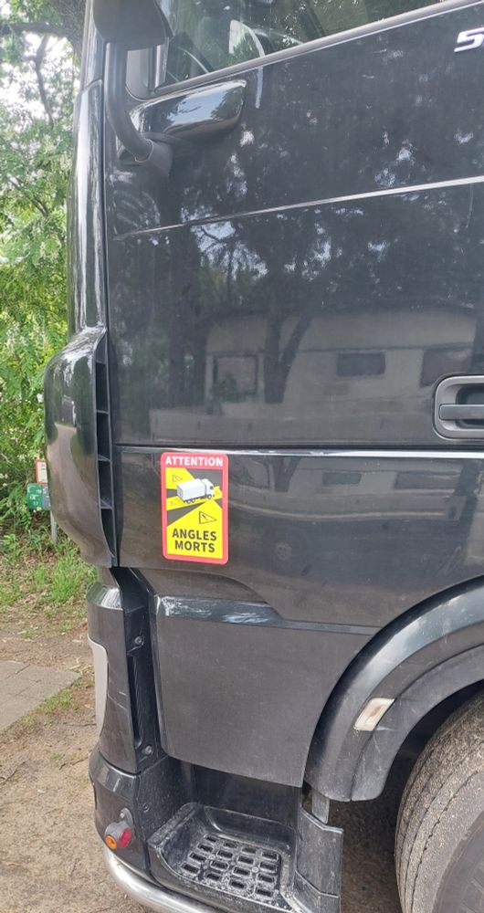 Auf der Fahrer:innen-Tür eines LKWs ist ein Schild: "Attention Angles Morts", auf dem die toten Winkel eines LKW schwarz markiert sind 