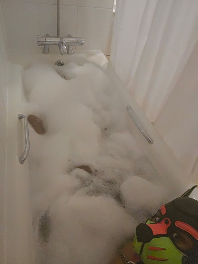 A bath filled to the brim with water, with a Wet Ass Puppo covered in bath bubbles!