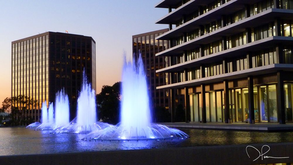 Four fountains in a line all spouting purple-lit geysers in the midst of a coppery sunset off the city architecture. 