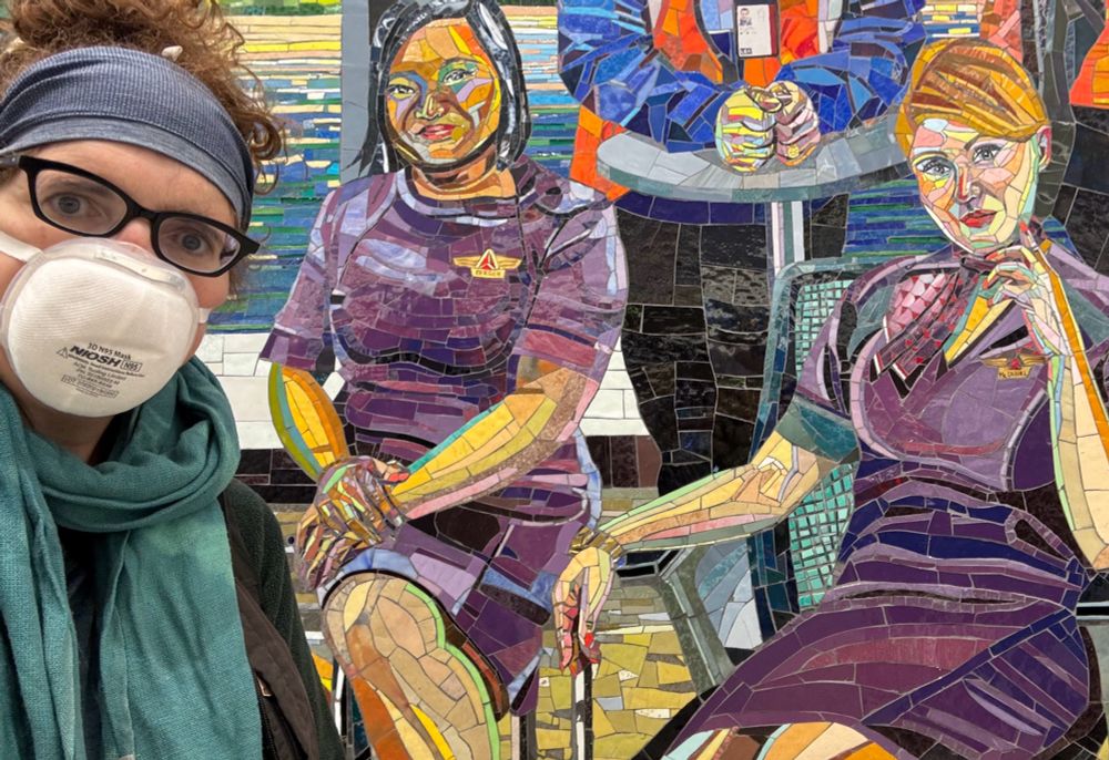 Selfie of me wearing a SoftSeal mask - also a blue headband, green scarf and dark vest -  in front of a mosaic picture in the Delta terminal at LaGuardia, which shows two Delta flight attendants in purple uniforms in casual chairs 