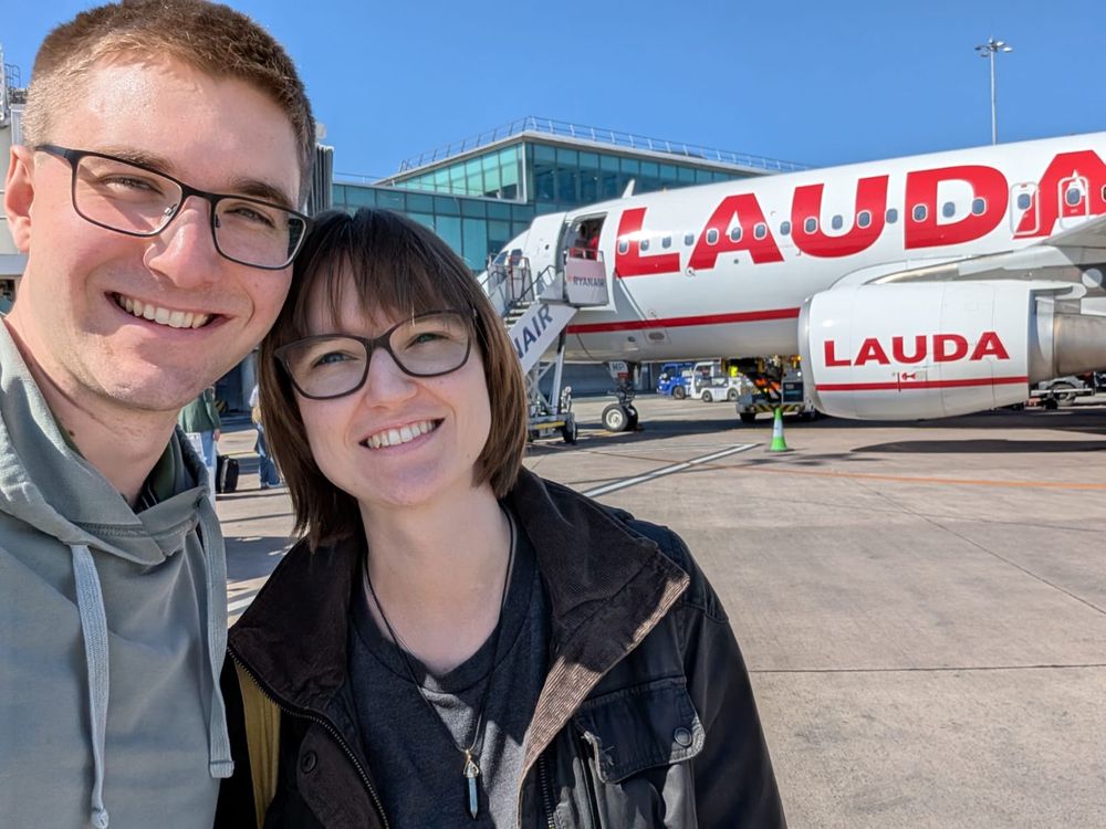 Catherine and her husband in front of a Lauda plane. The day is sunny and the plane is white with red capital text saying LAUDA and red design. Picture is at Manchester Airport