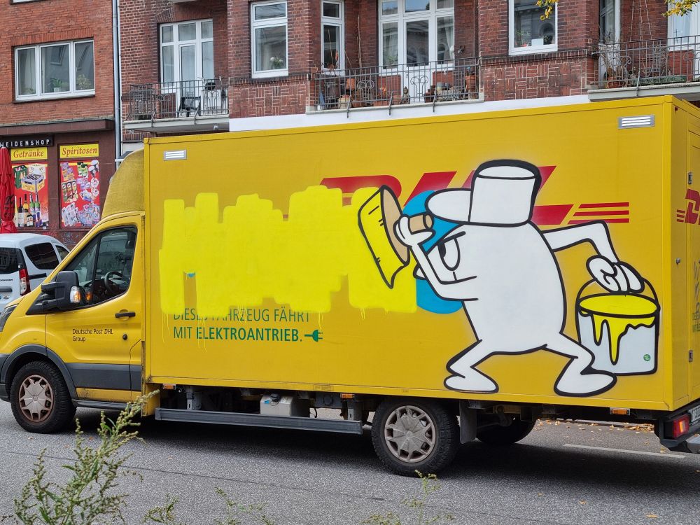 Ein DHL Wagen. Eine Figur aus dem Rosaroten Panter ist darauf gesprüht. Er mahlt die Schrift mit gelber Farbe über. 