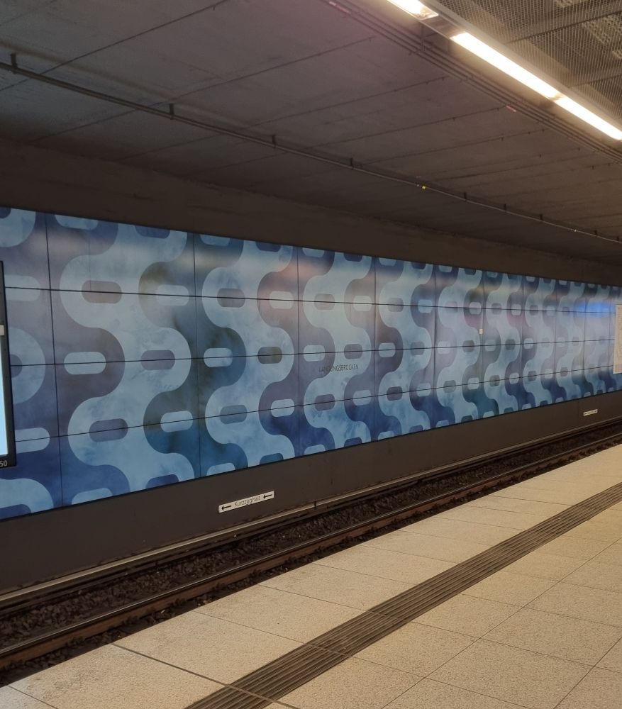 Bahnhof Landungsbrücken. Im Vordergrund der Bahnhof mit hell beigen Kachen die Wand hinter den Gleisen ist mit einem Hell und Dunkel- Blauen Wellenmustet gestaltet 