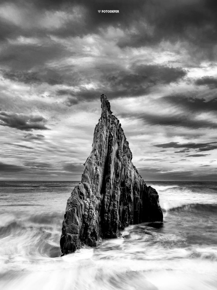 Una roca en el centro entre el mar y el cielo. Se erige como un centinela que destaca mucho sobre el fondo, lo que le da fuerza a la imagen.
Y la fotografía en blanco y negro le da un toque dramático y misterioso.