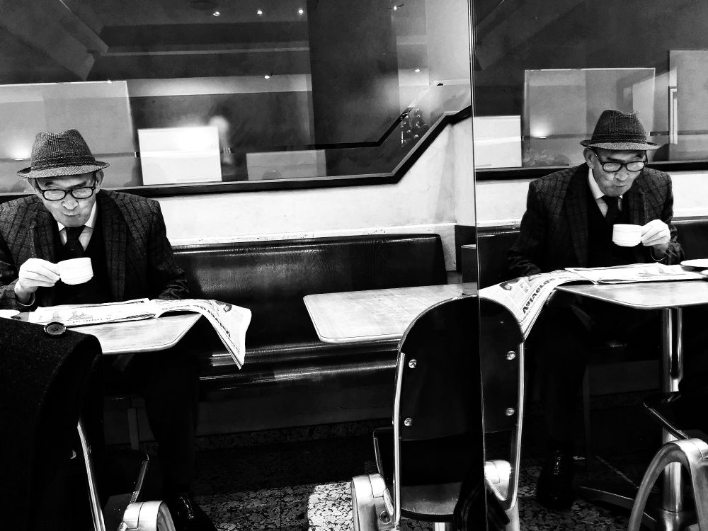 La imagen muestra a un hombre en un café, capturado en blanco y negro, lo que le da un aire atemporal y clásico. El hombre está vestido con un traje y un sombrero fedora, lo que sugiere una cierta elegancia o quizás un estilo anticuado. Está absorto en la lectura de un periódico y bebiendo de una taza, lo que sugiere un momento de soledad o contemplación.

Lo más notable de la foto es el reflejo del hombre en un espejo. Este reflejo duplica al hombre, creando una sensación de dualidad o de un "otro yo". El reflejo también introduce una especie de juego de perspectivas, ya que no estamos seguros de cuál es el "verdadero" hombre y cuál es el reflejo.

"La otra mesa" es un título evocador que invita a la reflexión sobre la naturaleza de la realidad, la identidad y la percepción. La foto, con su juego de reflejos y su atmósfera melancólica, complementa perfectamente este título, creando una sensación de misterio y ambigüedad.
