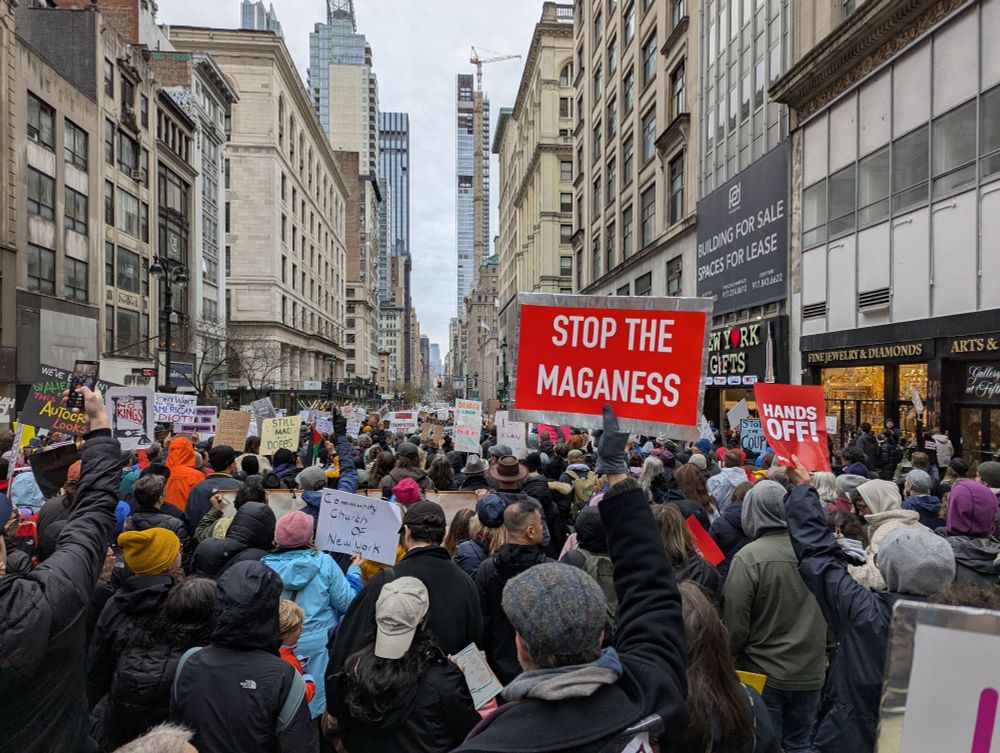 Crowd of protestors in NYC on Fifth Avenue. The street is completely filled. A red sign says "Stop the MAGANESS."