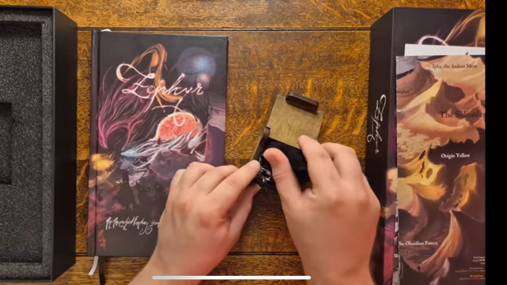 A screenshot from Cozy RPG Reviews’s Zephyr video, the Zephyr book is slightly off-center to the left, and in the center is a black box being tinkered with by the YouTuber’s hands.

Left of both is an upside down big box showing foam inside, and on the far right of the image is the other half of the box, showing the art of the book for ZephyrRPG, and above the far-right box is the map for the game, folded up.

All of this is on a brown wooden table.