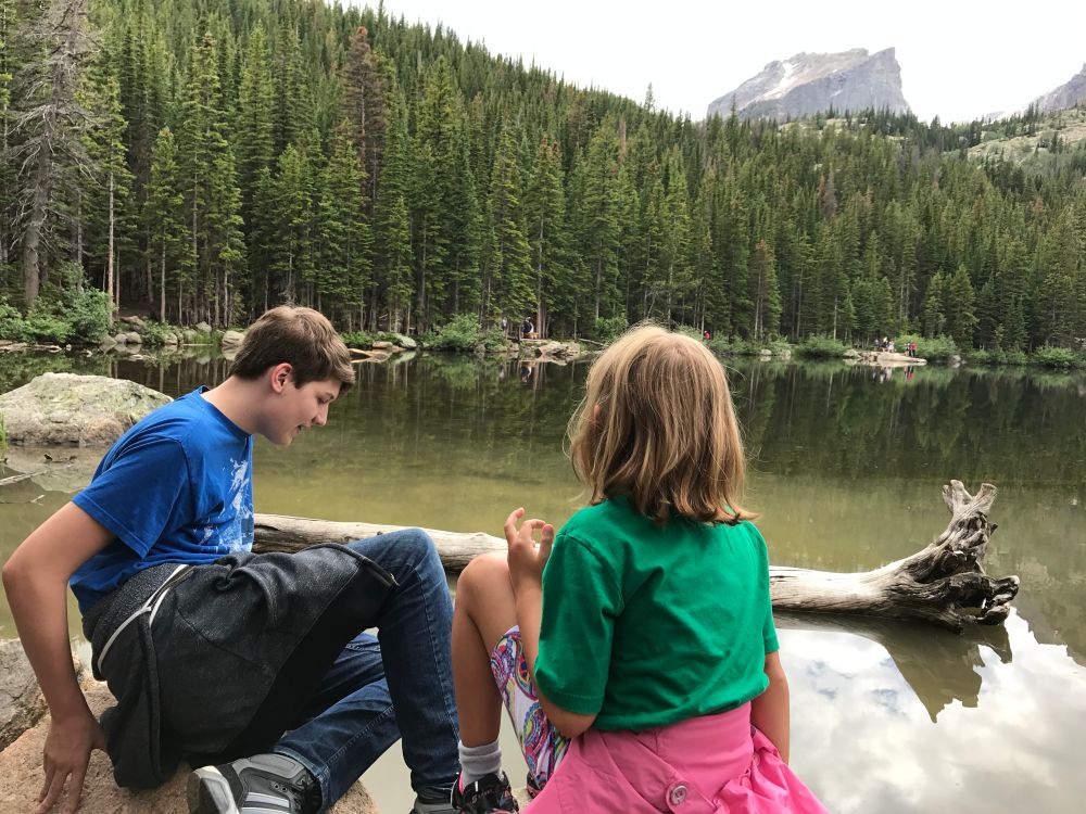 A photo of two kids by a lake, pine trees and a mountain peak in the distance, Rocky Mountain National park. 