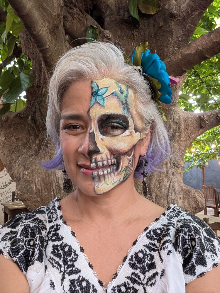Keynote speaker in Day of the Dead-inspired makeup where only half of her face depicts a skull with a background that intertwines agricultural fields, a corn crop, a hummingbird and a flower