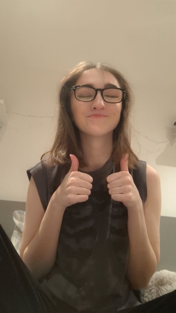 A photo of Chlo, they are sat cross legged on their bed with both thumbs up and their eyes closed, they are tired from staying up until 3am to fight a boss in a video game.
