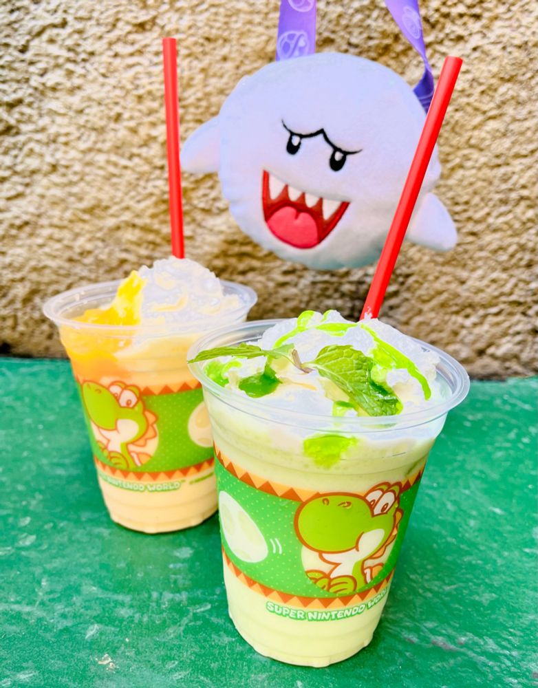 ユニバのニンテンドーワールド内、ヨッシーのドリンクの写真　手前に緑のメロン味ドリンク、奥に黄色のマンゴー味ドリンク、ドリンクのコップにヨッシーの絵が書いてある
上にテレサのぬいぐるみ