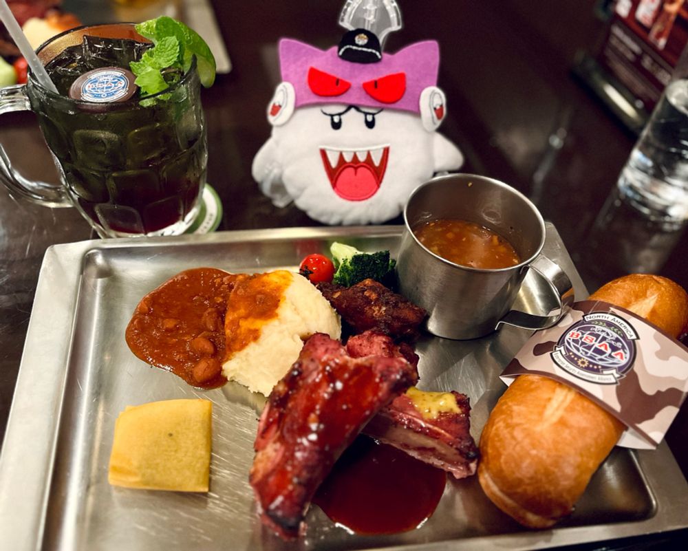 レストランでのテーブルの上の写真　手前に銀の四角いおぼん　お盆の上に肉塊とサンドイッチとスープが乗っている　左奥にコーラinジョッキ　奥中央にテレサちゃんのぬいぐるみ