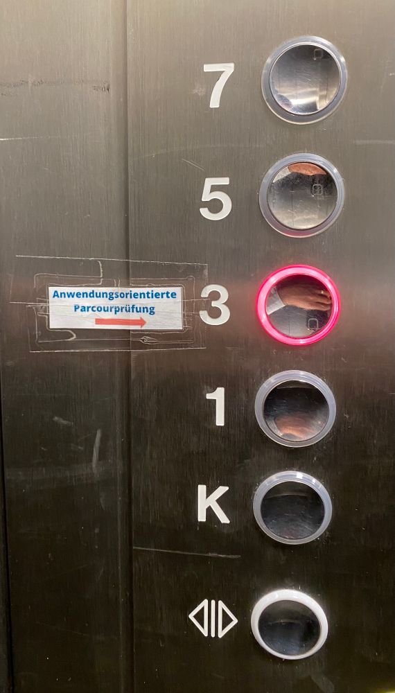 Aufnahme aus einem Fahrstuhl, an den Etagenknöpfen klebt ein Schild, das auf den Ort der Prüfung hinweist. 