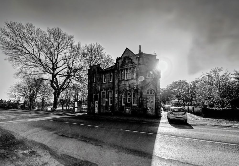 An abandoned building, once a silent shell, is now a vibrant canvas for graffiti, standing alone as the sun crests the top right of its roof, casting some lens flare.The graffiti is a testament to urban creativity.
To the left, a joyful looking leafless tree adds contrast to the scene. Its dark, skeletal branches, silhouetted against the pale morning sky, stretch out with an almost cheerful energy, contributing to a delicate, natural beauty that makes the otherwise desolate setting look pretty. The early morning light infuses the entire picture with a warm hue, creating a striking image of decay and renewal, where human artifice meets the resilient spirit of nature.