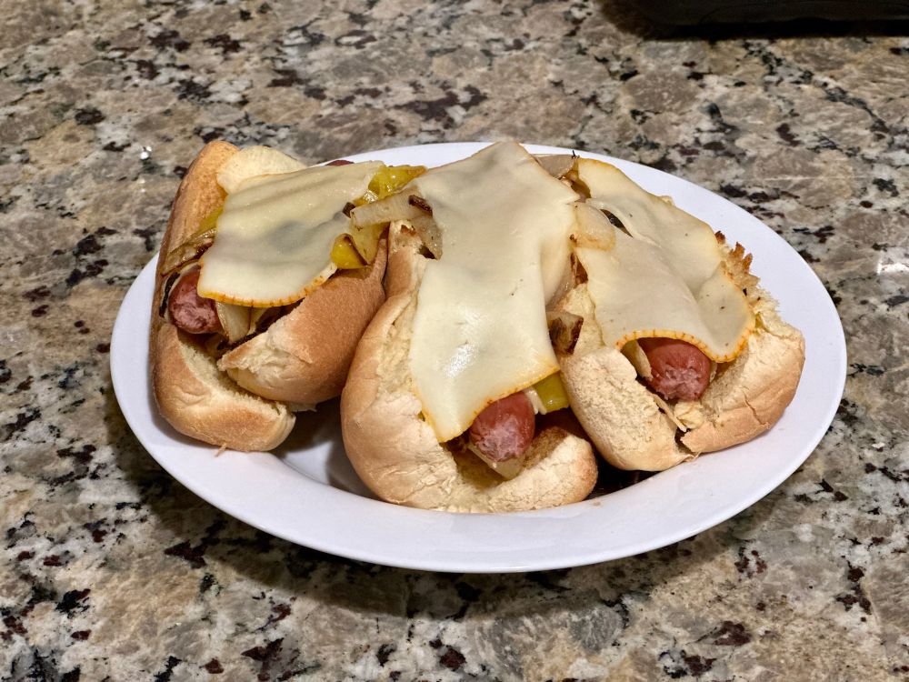 Three hotted dogs on a plate, with toasted buns, chips underneath, grilled peppers/onions/garlic, Dijon, Muenster on top.