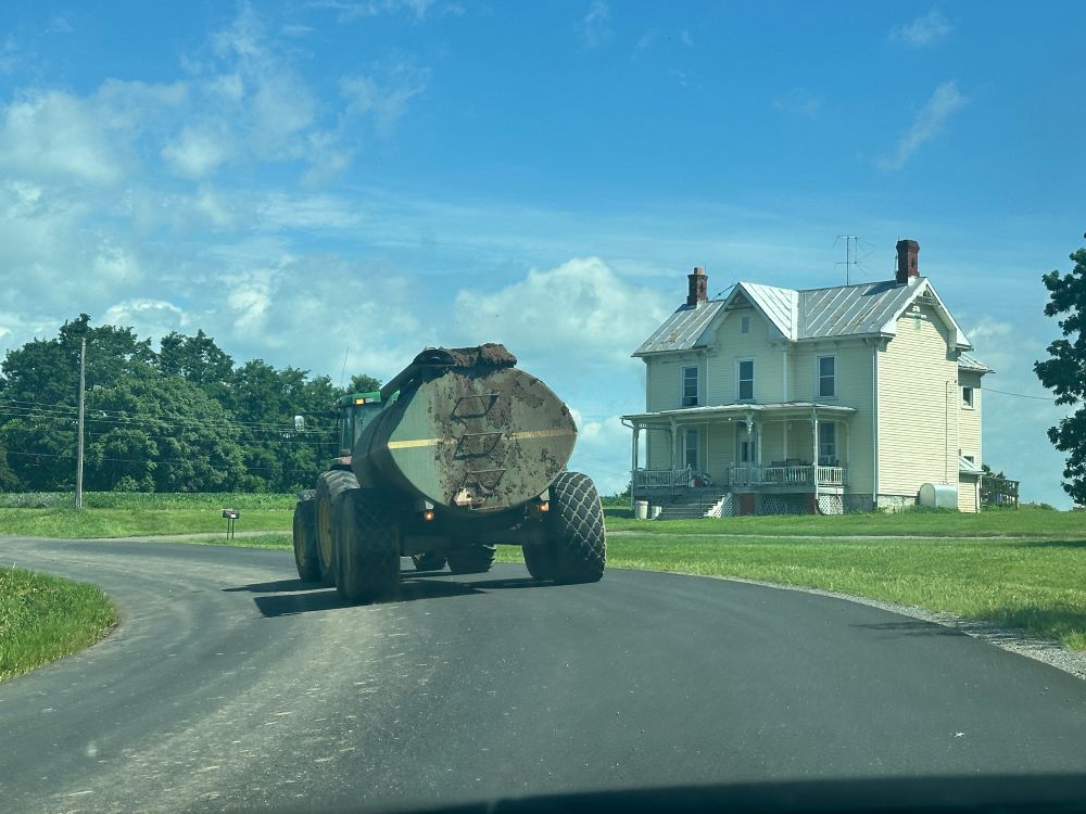 Sewage truck going down road with white farmhouse adjacent. 