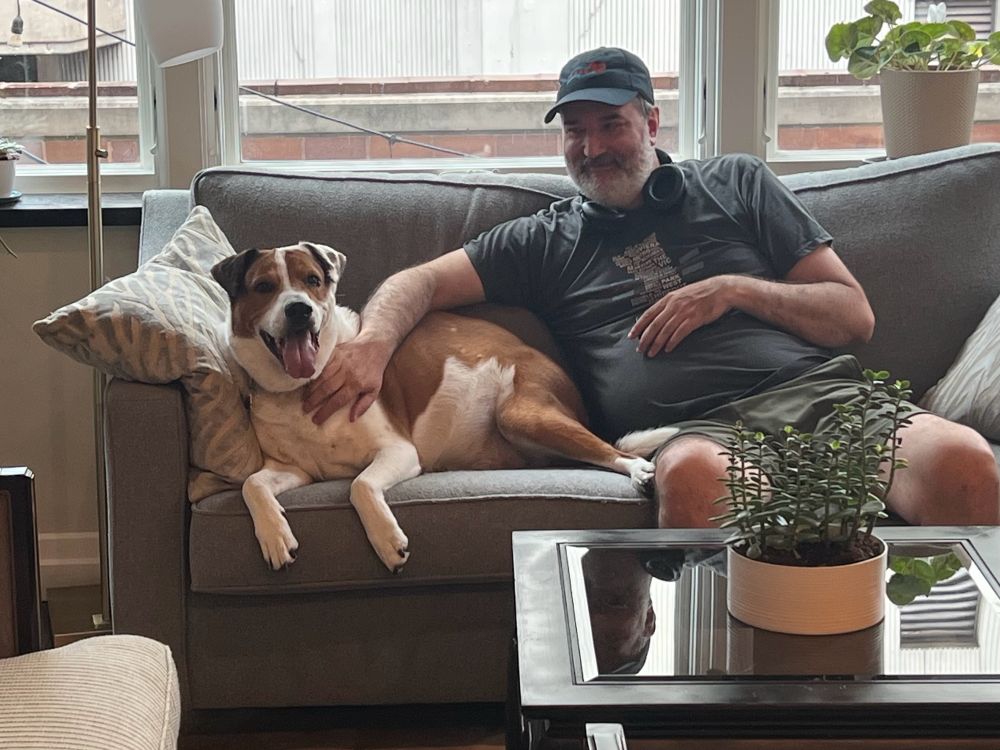 Man smiling while sitting on a couch with a large, happy brown and white dog 
(David and Dozer)