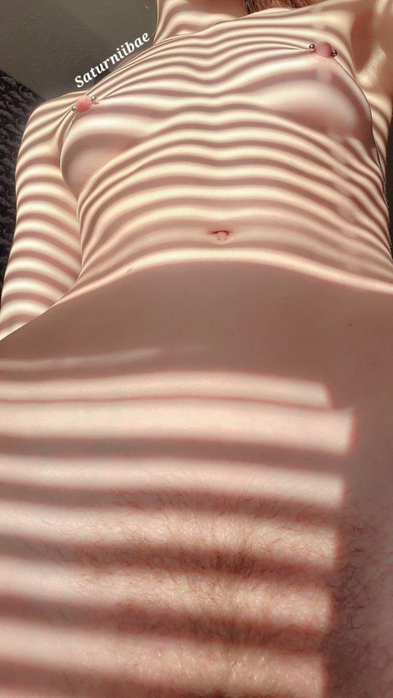 A photo capture from a low angle of Saturn's nude figure, illuminated by the sunlight. Patterns of stark shadows contrast the golden light falling on her skin, drawing the viewer's eye across the landscape of her body. Accents of muscles can be seen lining her lithe abdomen, round navel placed delicately in the center of her stomach. Silver piercings decorate her perky pink nipples. Hair like gold delicately grows on her pelvis, leading toward the true treasure below.