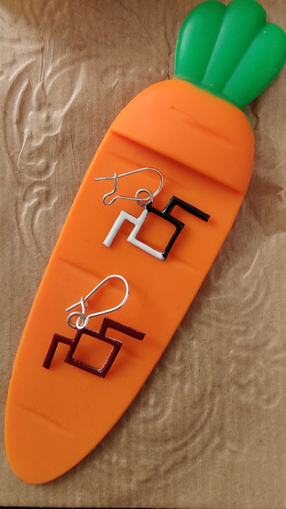 Pendientes sobre una zanahoria de plástico en 2D. Los pendientes son de Nebel Studio, el modelo Las lechuzas no son lo que parecen, inspirados en la serie Twin Peaks. Están esmaltados, representando el símbolo   de los espíritus en dos colores: por un lado negro y blanco y por el otro negro. 