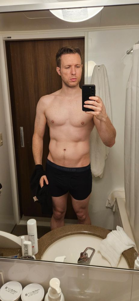 A shirtless man taking a bathroom mirror selfie