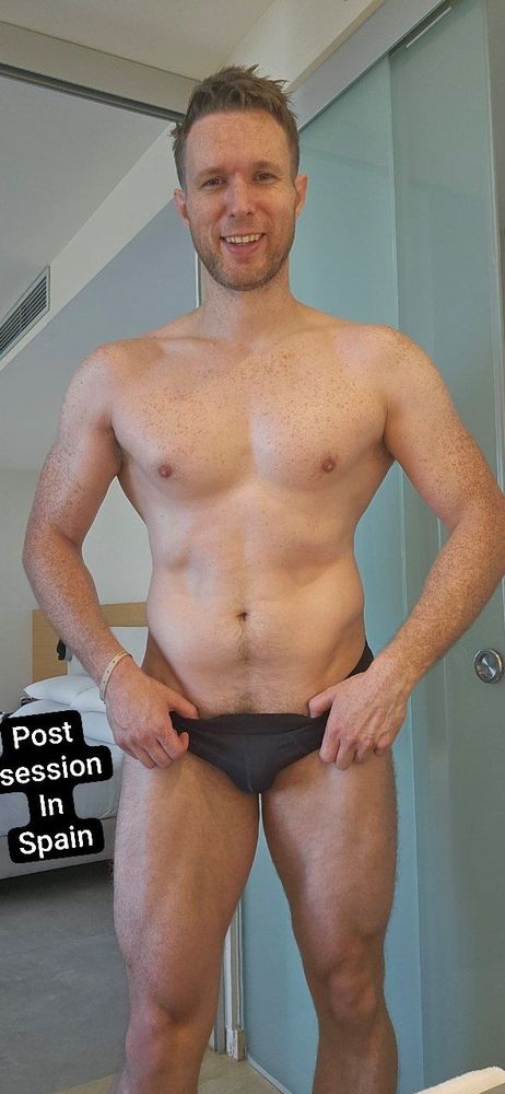 A man poses in a pair of small briefs in a way that emphasises body progress in a bathroom with text that reads "post session in Spain"