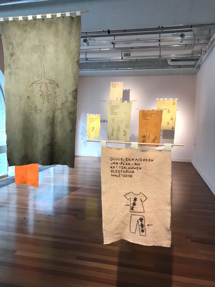 Wollen doeken hangen in de Witte Zaal bij de Brakke Grond. Gekleurd met natuurlijke kleurstoffen van planten uit de Lage Landen. Op de doeken zijn oude Nederlandstalige namen van de planten geborduurd.