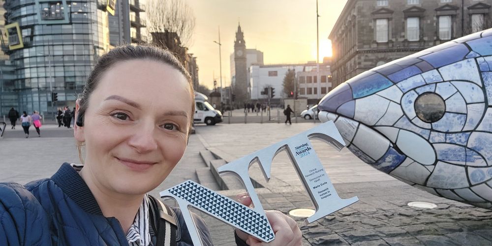 A woman takes a selfie in Belfast, holding a Nursing Times Award 2024 for Clinical Research Nursing, which is inscribed with "Marie Curie Research Nurses in hospices". She wears a navy quilted jacket and stands beside the iconic "Big Fish" mosaic. The Albert Memorial Clock and a modern glass building are visible in the background, with the setting sun casting a warm glow.