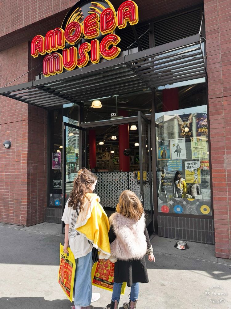 Two little girls standing, back to back, facing the entrance to Amoeba Music on Hollywood Blvd. They each hold a bag from the store containing an LP.