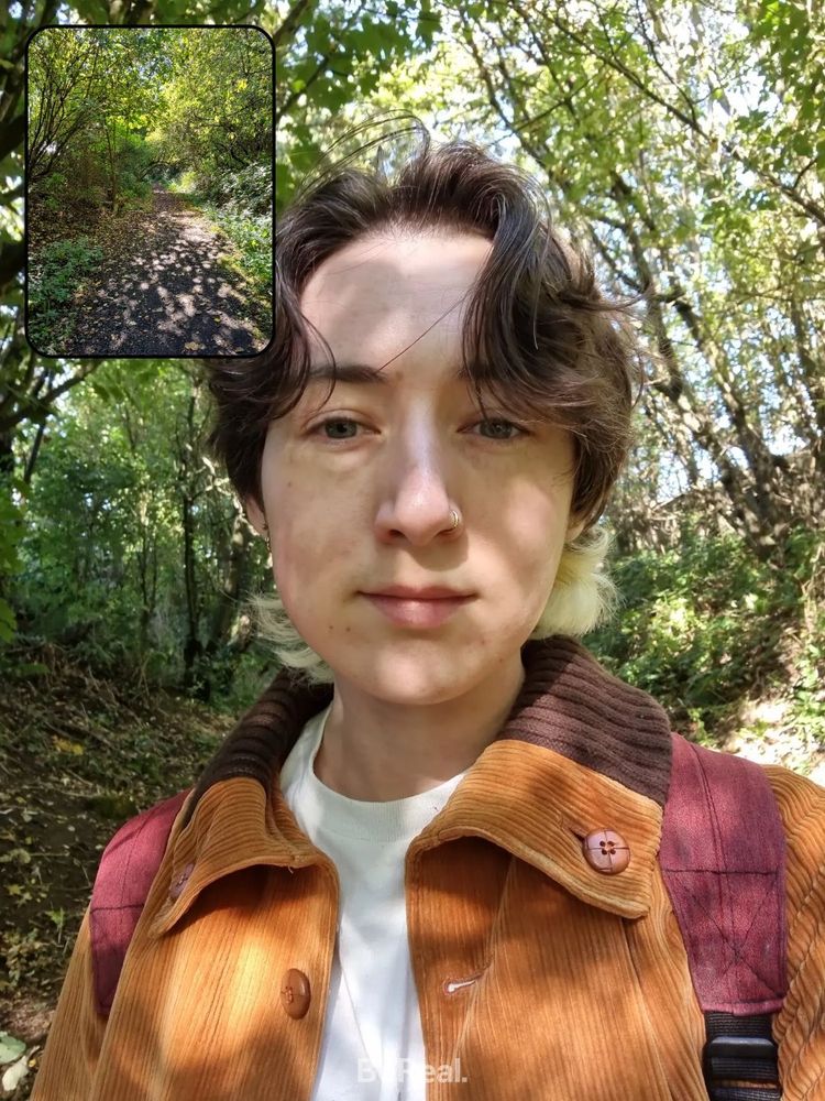 A photo of Robin, who has pale skin and dark mid-length hair bleached underneath. He is wearing an orange corduroy jacket and walking through the woods.