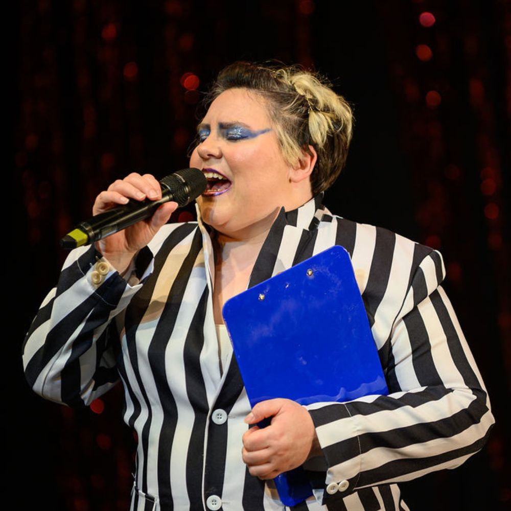 A stage performer wears a black and white striped blaze, they have blue eyeshadow that sparkles with a blue clipboard tucked under their arm. They sing into a microphone, their eyes are closed.