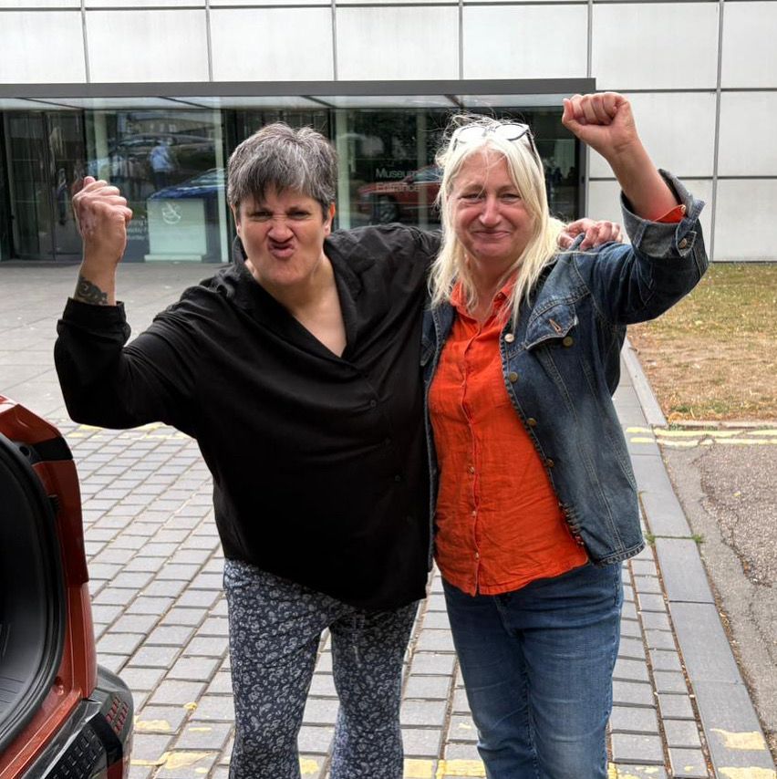 Dolly Sen and DaDa CEO Zoe stand in a street next to a car with its boot open. They are raising a fist and smiling