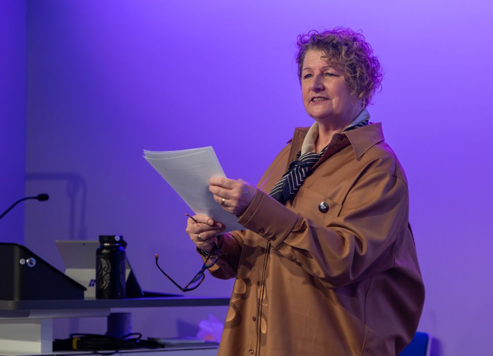  Kaite O'Reily is pictured delivering her lecture for DaDaFest at the Rushton Lecture. She is holding a few pieces of paper, at a podium, looking upward confidently at the audience. She is stood in front of a wall that is lit with purple lighting. She wears a beige shirt with a blue striped tie. 