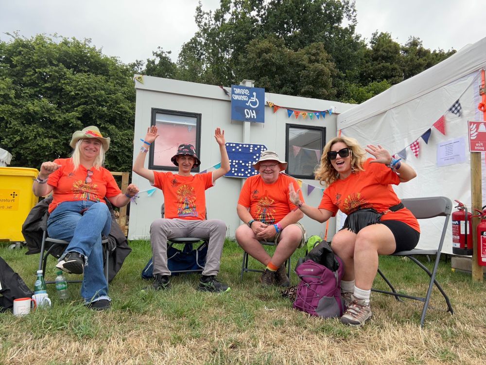 Four people wear orange festival T-shirts. They are seated in camping chairs with porta cabins behind them in a field. There are blue signs with a wheelchair symbol on behind them. 