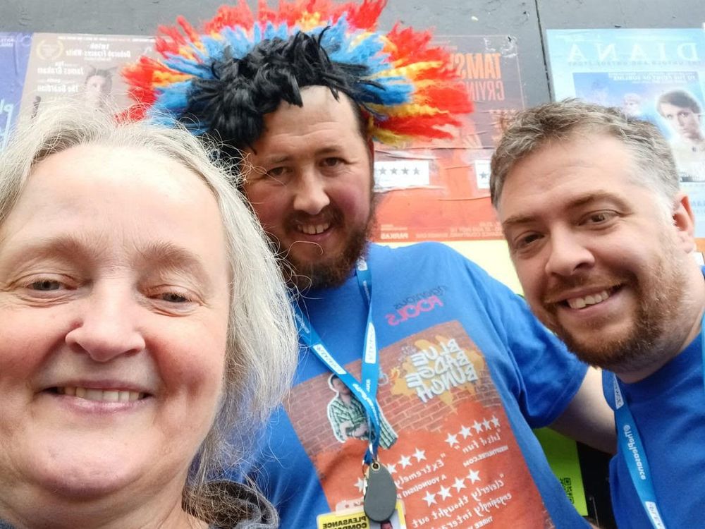 An image of three people smiling brightly, it's mostly an image of their smiling faces, the person in the middle of the image has a bold blue t-shirt on featuring a graphic image, which appears to be an advertisement for a show. They are all at the Edinburgh Fringe Festival. They are stood in front of a green wall which has blurry posters adorned all over it. The person in the middle and the right of the image have a blue lanyard on. 