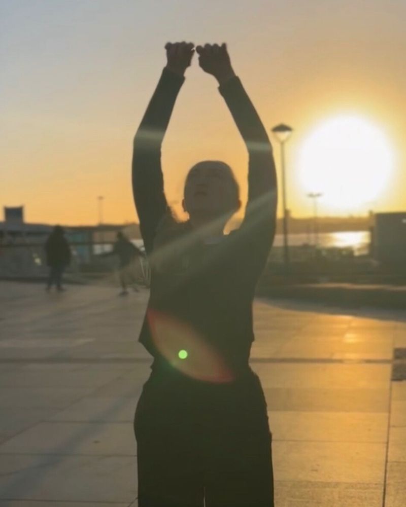 An image of Amy Milson, a dancer, with arms outstretched to the sky, performing a dance piece outdoors. The sky is glowing orange, the sun very present in the background of the image, setting in the horizon. In the background, the water at Liverpool docks glimmers as it reflects the sunshine. Amy is partly a silhouette due to the volume of light from behind her. 