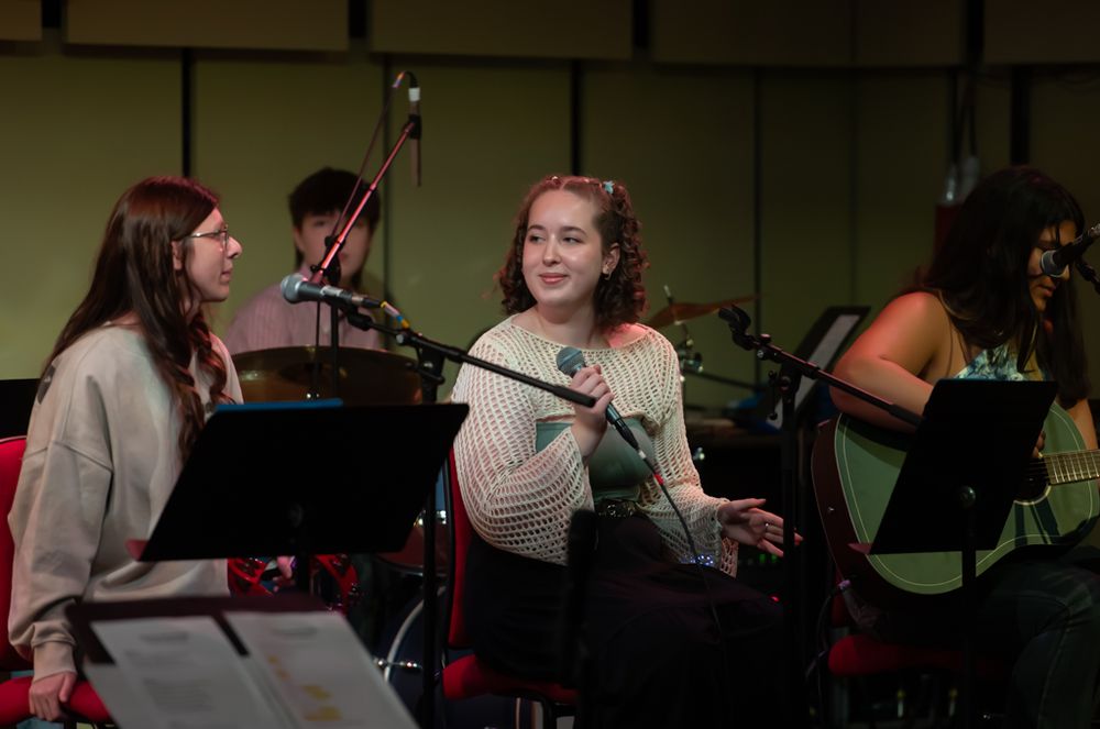A girl with curly shoulder length hair partially tied up is holding a microphone sitting next to one girl playing the guitar and another with long hair and glasses sat at a mic. Behind is another teenager obscured.
They are in stage lighting.