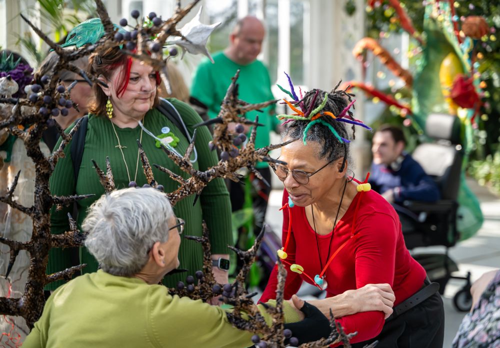 An image of Janet Price and Faith Bebbington at the launch of Pimp My Wheelchair. The two artists hold one anothers arms, and look at one another with love and adoration. Faith wears a green jumper, Faith wears a red one. Thorns from installations are present in the image. Others are pictured in the background, blurry. 