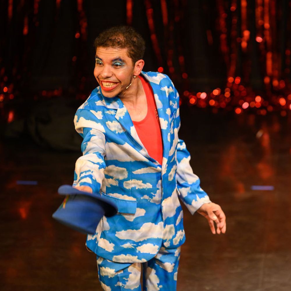 A cheerful performer dressed in a bright blue suit patterned with white clouds smiles widely and extends a blue hat toward the audience. They wear vibrant blue eye makeup and red lipstick, standing in front of a sparkling red curtain backdrop.