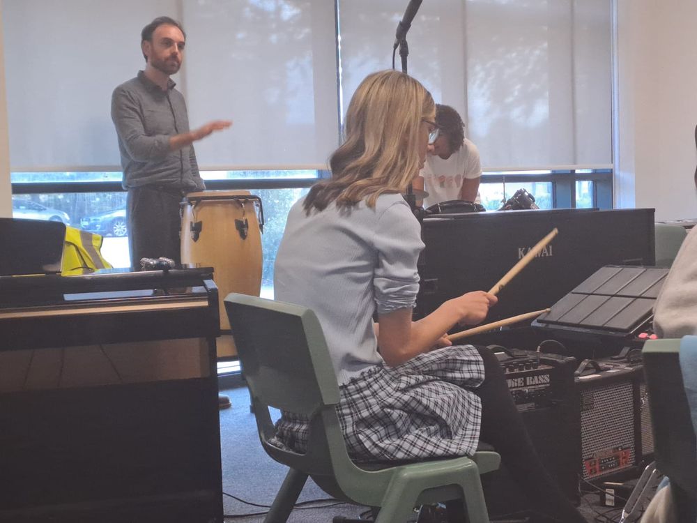 A man is standing at the back of a room banging on a drum. A young person has their back to camera at the front using drum sticks to tap on electronic pads.