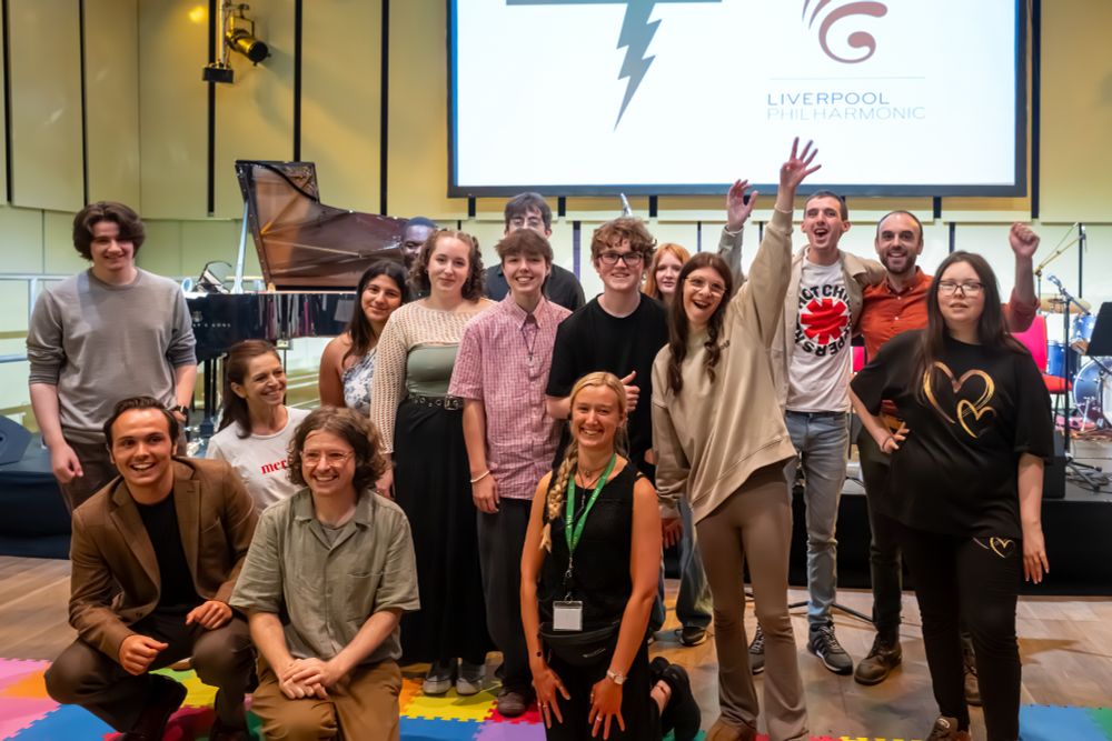 A group of disabled musicians are gathered in a group in a music performance space. Instruments are behind them including a grand piano. There is a large screen with logos on. Some are seated and some are standing. Some have hands raised in excitement and they are smiling.