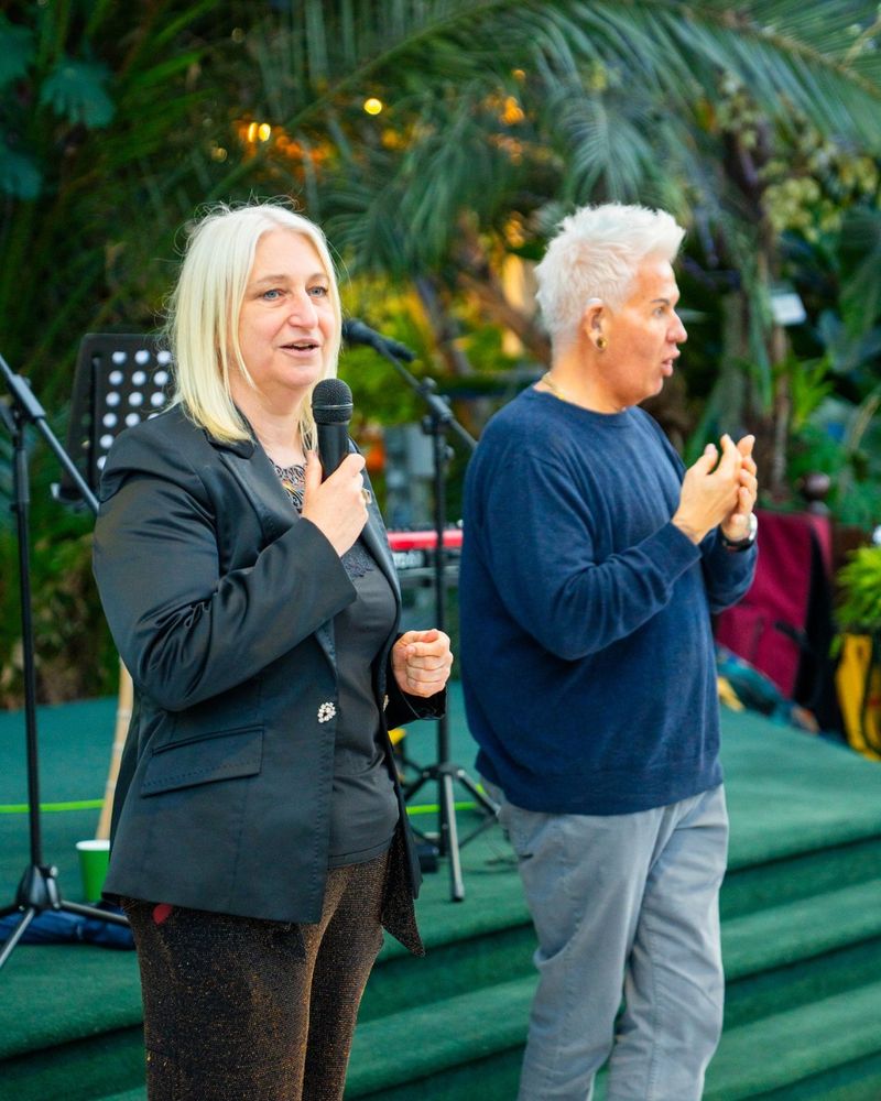 DaDa CEO Zoe Partington dressed in a smart black jacket and trousers holds a mic. Beside her is a BSL interpreter. Behind them are palm trees and foliage.