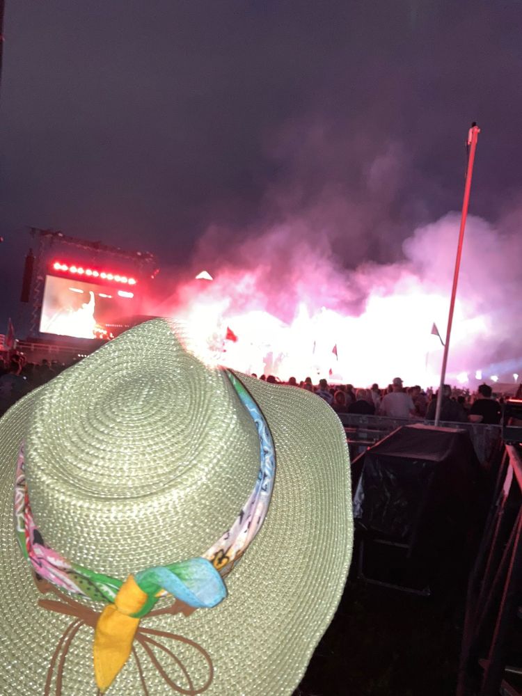 DaDa CEO Zoe Partington wears a straw hat facing away from the camera towards a festival stage which is in the distance with a large blurred screen. It is night time. There is smoke and bright lights in front of the stage hovering over a large festival crowd. 
