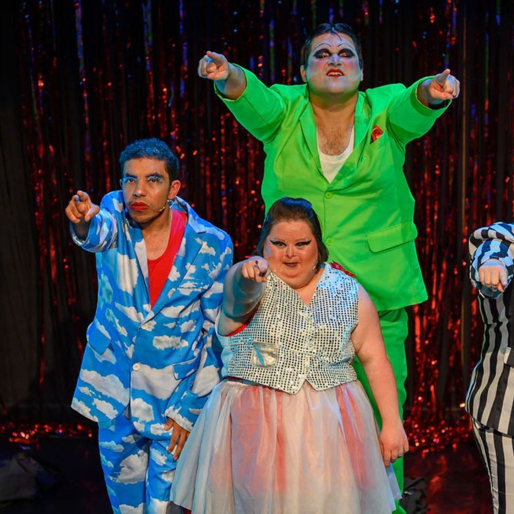 Three performers stand together on stage, dramatically pointing toward the camera. The front performer wears a silver sequin vest and a pink tulle skirt. Behind them, a performer in a green suit and another in a cloud-patterned blue suit strike a strong pose, all with bold, theatrical makeup.
