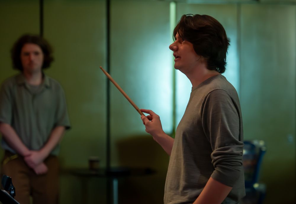 A young man with wavy dark hair just above the shoulder is holding a stick and conducting.