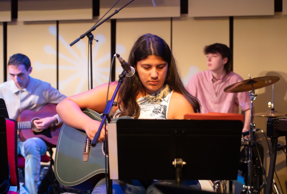 Image description
A teenage girl with long dark hair is sat at a music stand playing the guitar. Just visible over her should is a boy playing the guitar and another at a drumkit