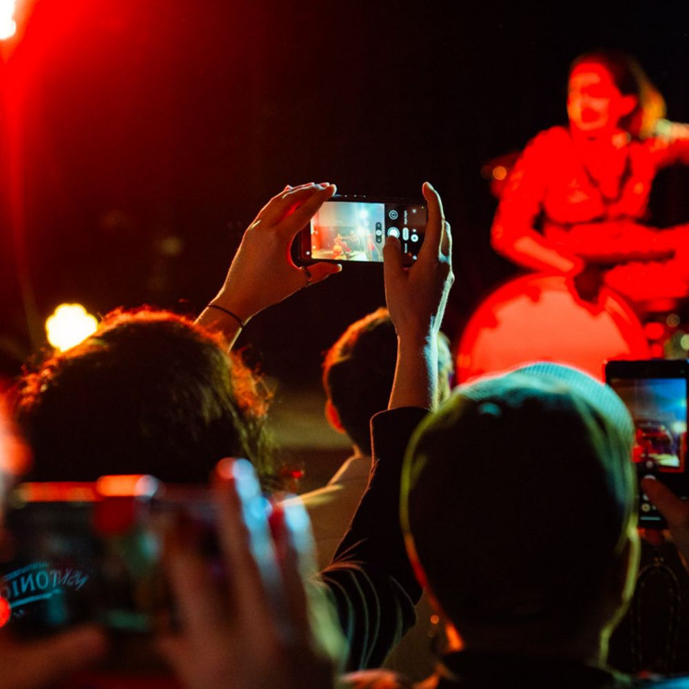 At the foreground, someone is pictured holding a phone in the air, filming the action on stage. A lively audience pictured from behind. The background is of performers on stage, blurry, red light.