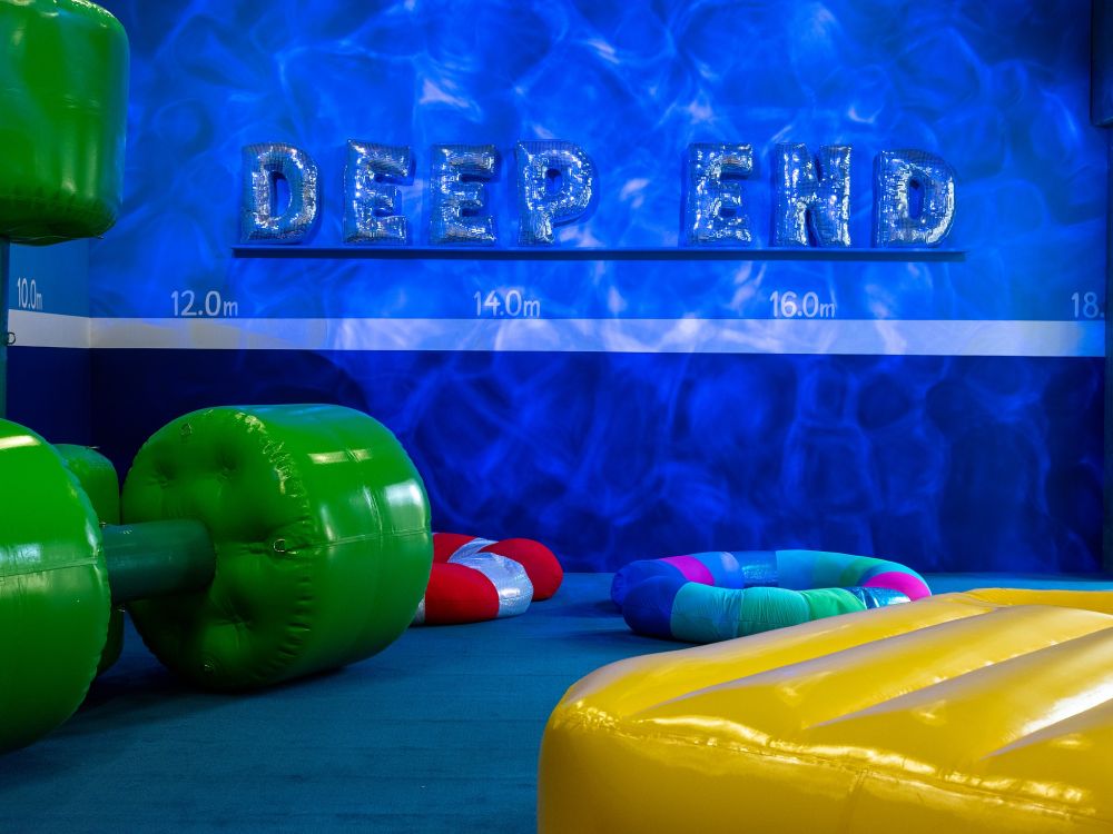 A bright blue wall carries inflated letters reading 'Deep End'. Beneath is a yellow inflatable Lilo on the floor next to green inflatable dumbbells.