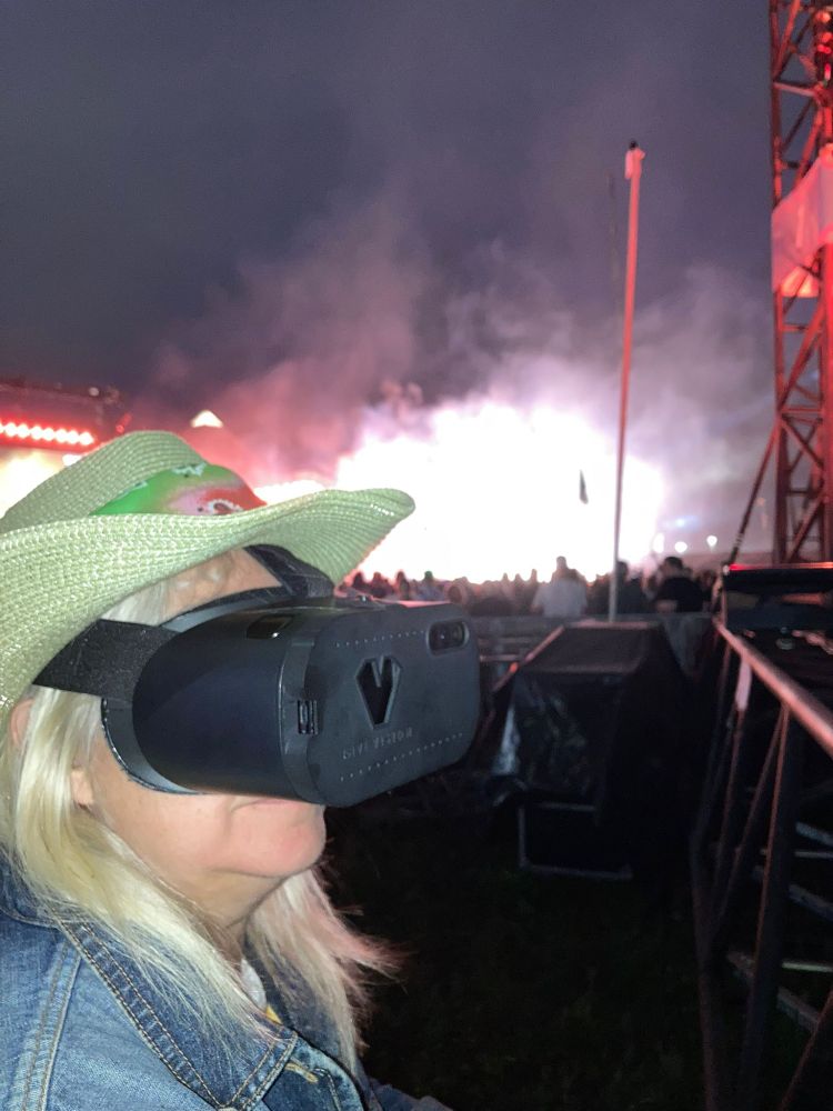 DaDa CEO Zoe Partington stands wearing a straw hat and denim jacket. She has a VR headset on and a night time festival crowd can be seen behind her.