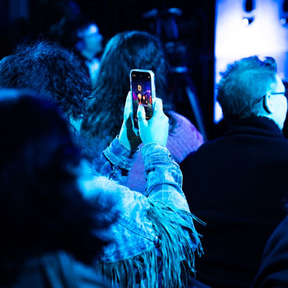 An image taken from a busy crowd, we are in and amongst it. The audience is an audience from a drag cabaret. The light is blue and vivid. Someone is filming the activity on stage, featured is their hands holding a phone, this is the primary focus of the image. There are backs of heads featured in the image, blurry and blue, behind. 