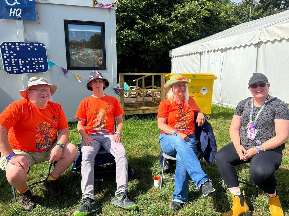 Two men and a woman wear orange festival T-shirts. Another woman is dressed in leggings and a grey T-shirt. They are seated in camping chairs with porta cabins behind them